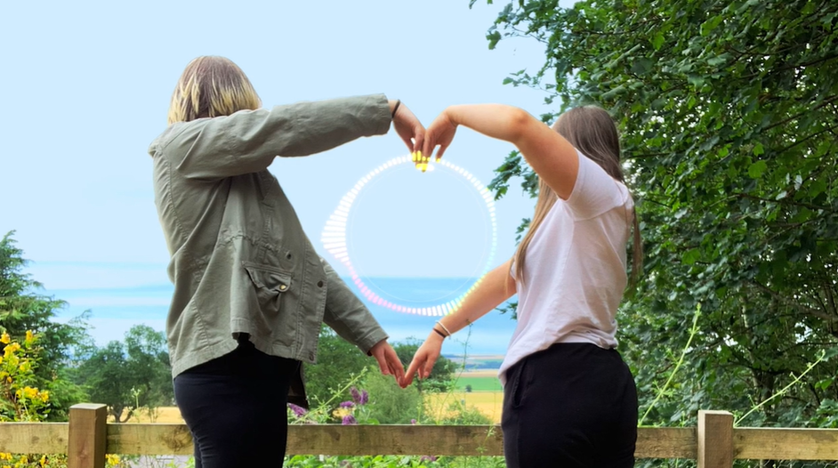 Two people making a love heart shape using their arms, looking away from the camera to the blue sky in the background.